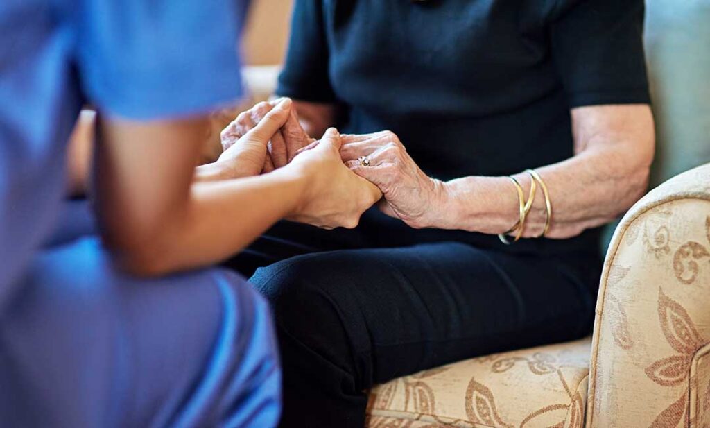 Elderly couple sitting together in a bright living room, one reading while the other receives gentle help from a caregiver.